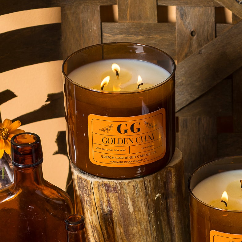 Two amber glass jar candles with orange labels are displayed on a rustic wooden shelf. One candle sits on a wood block and the other on a teal base, with small amber bottles, flowers, and jars in the background.