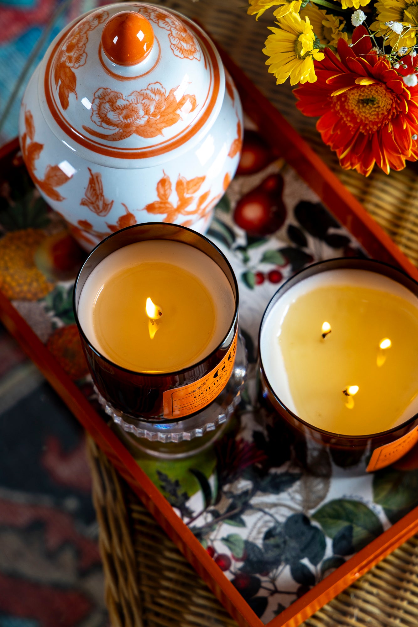 Two lit candles on a decorative tray with a floral-patterned vase and flowers in the background.