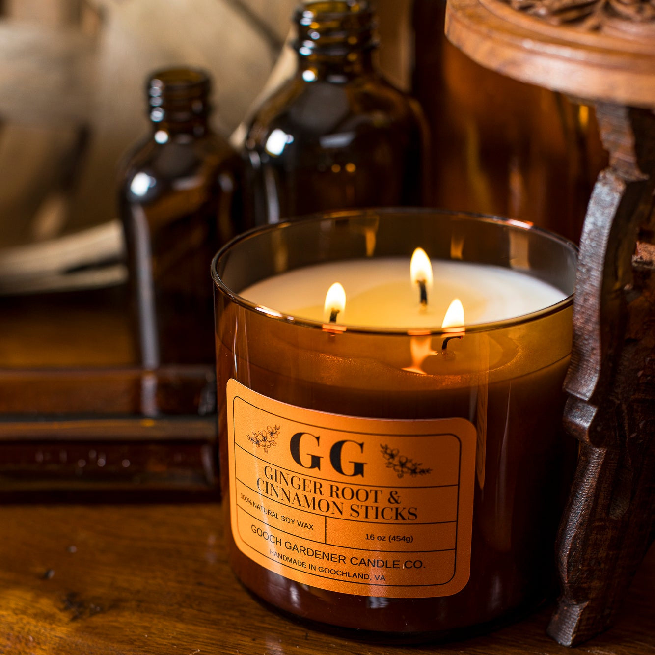 A lit amber glass jar candle with three flames sits on a wooden cabinet. The candle label reads &