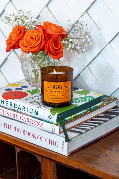 Candle with 'GG' branding on a stack of books with orange flowers in the background