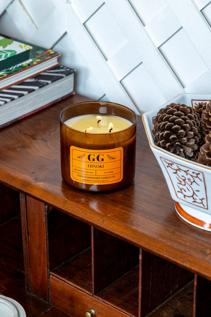 Candle with an orange label on a wooden surface next to books and a decorative pot with pinecones.