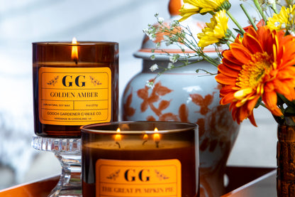 Two lit candles with 'GG' labels on a decorative surface with flowers.